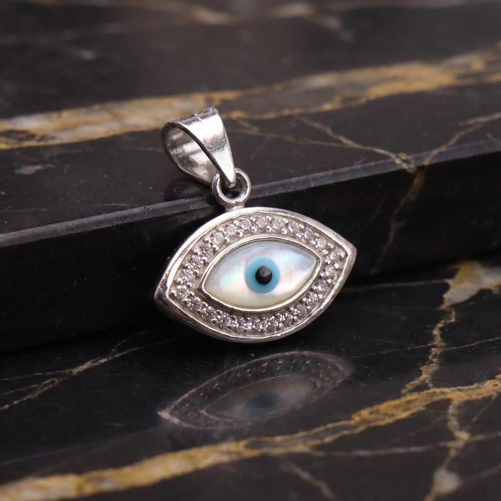 A sterling silver evil eye pendant with a white mother-of-pearl background and a blue iris, displayed on a black and gold marble surface.