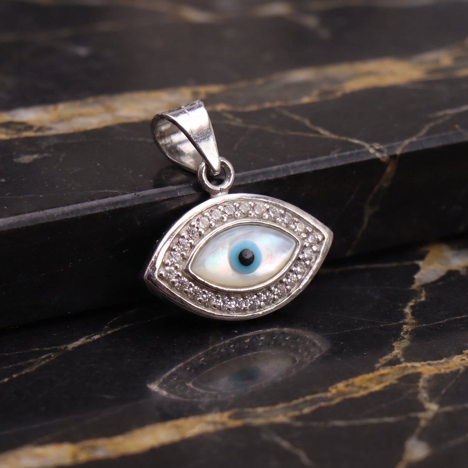 A sterling silver evil eye pendant with a white mother-of-pearl background and a blue iris, displayed on a black and gold marble surface.