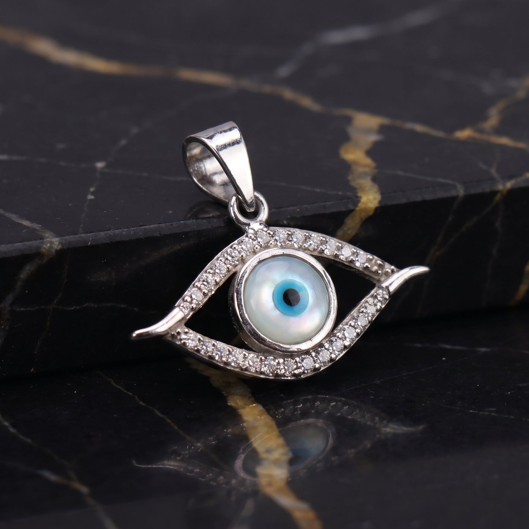 A sterling silver evil eye pendant with a blue and white glass eye in the center, surrounded by small clear crystals, resting on a black marble surface with gold veining.