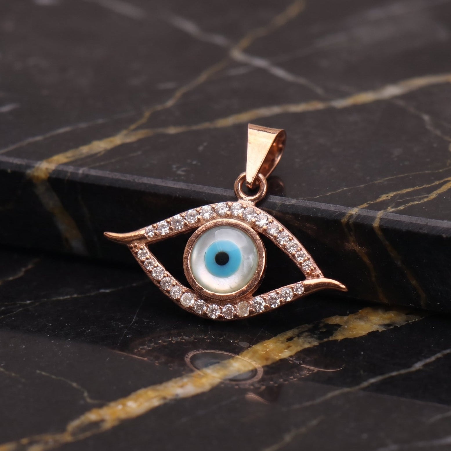 A rose gold evil eye pendant with a blue and white center, surrounded by small clear crystals, resting on a black and gold marble surface.