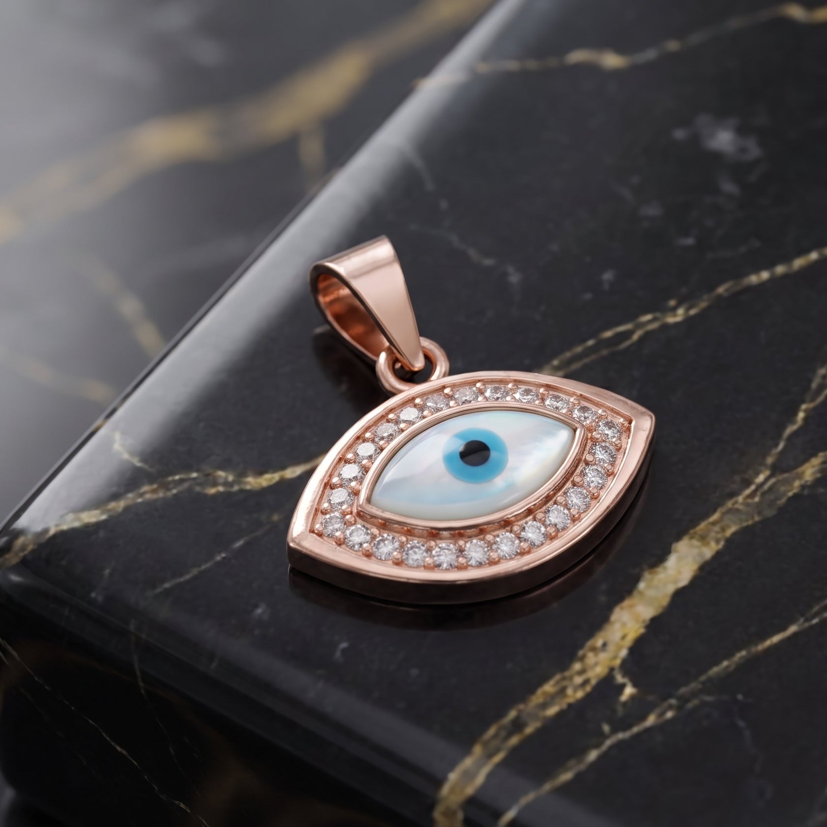 A rose gold evil eye pendant with a light blue and black eye design, surrounded by small clear stones, resting on a black marble surface.