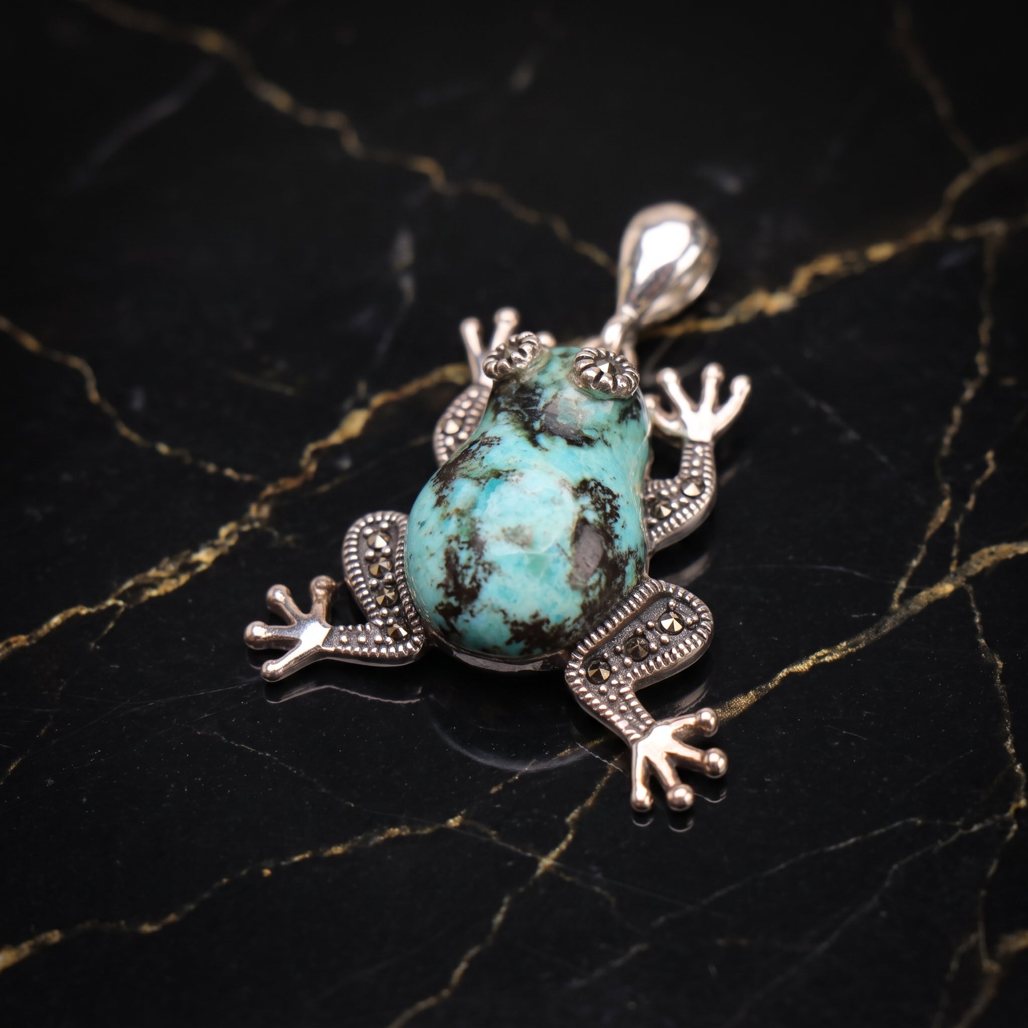 A frog-shaped pendant featuring a turquoise-colored stone body and silver-toned metal limbs and head, displayed on a black marble surface with gold veining.