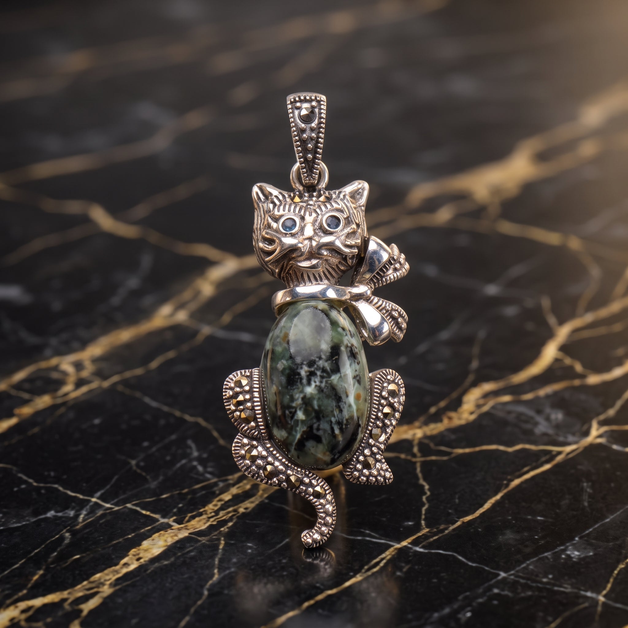 A silver cat-shaped pendant featuring a large green and black marbled stone belly and detailed silver accents, displayed on a black marble surface with gold veining.