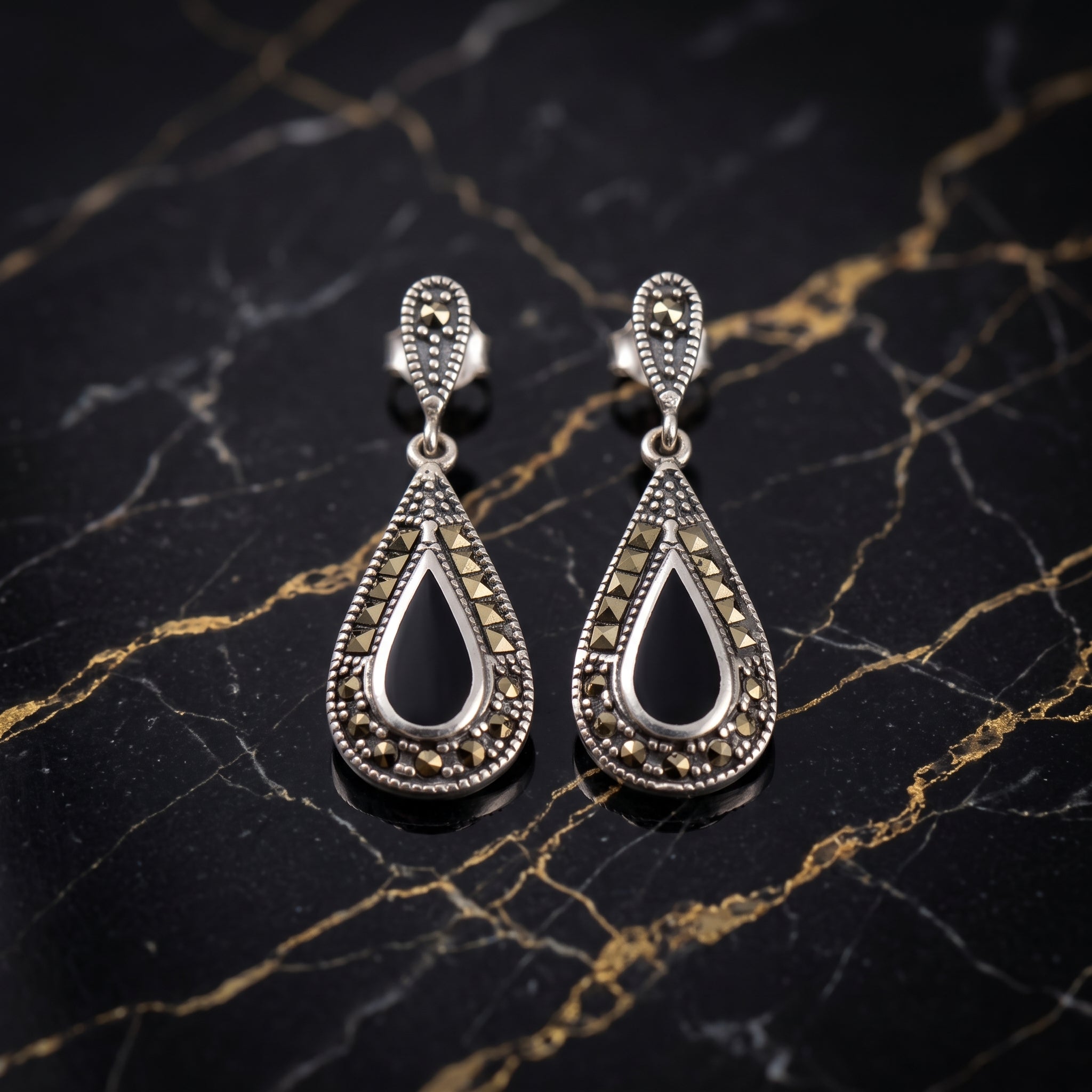 A pair of silver teardrop drop earrings with ornate filigree detailing and small marquise-cut stones, displayed on a black and gold veined marble surface.