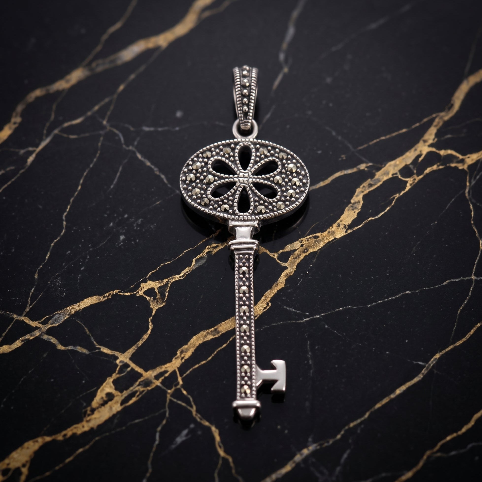 A vintage-style key pendant made of silver-toned metal with pavé-set clear stones, displayed on a black and gold veined marble surface. Marcasite Stones