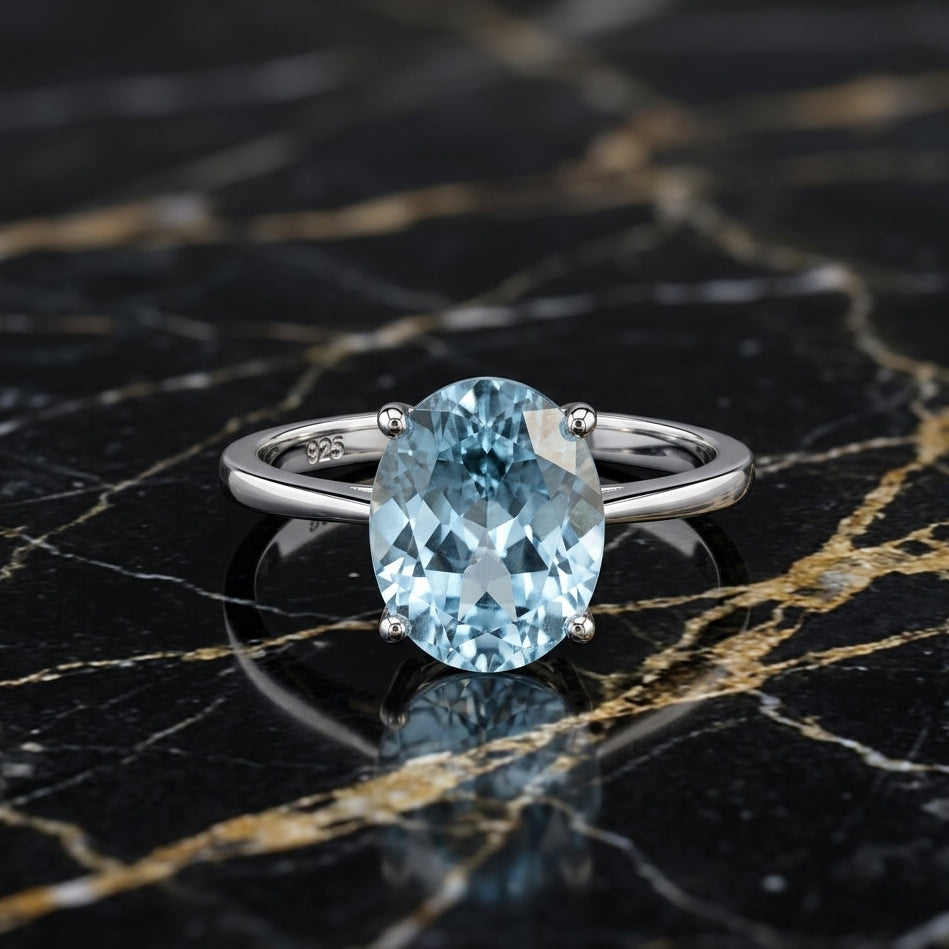 An oval-cut light blue gemstone ring set in a silver-colored band, displayed on a black marble surface with gold veining.