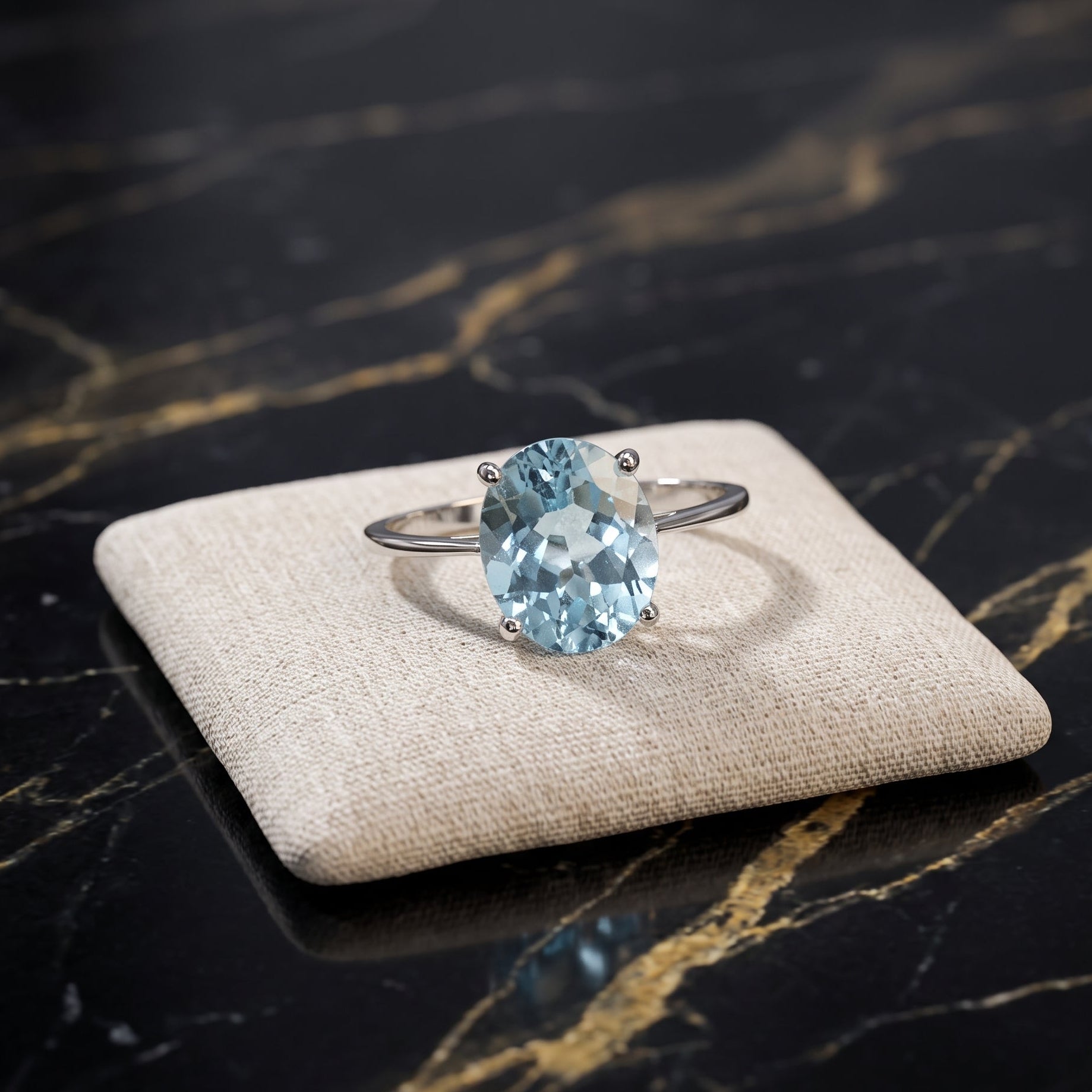 A silver ring with a large oval light blue gemstone, displayed on a beige fabric cushion over a black marble surface with gold veining. Blue Topaz