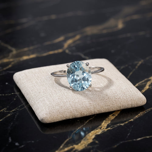 A silver ring with a large oval light blue gemstone, displayed on a beige fabric cushion over a black marble surface with gold veining. Blue Topaz