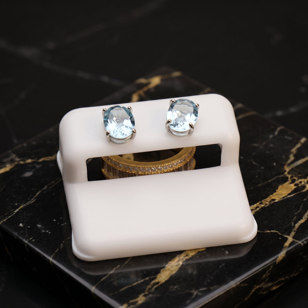 A pair of oval stud earrings with light blue gemstones in silver settings, displayed on a white stand over a black marble surface with gold veining.