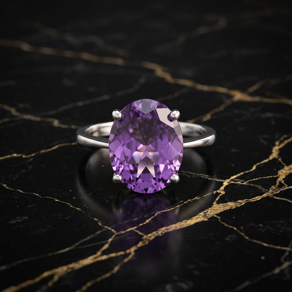 An oval-cut purple amethyst ring in a silver four-prong setting, displayed on a black marble surface with gold veining.