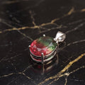 A silver-toned oval pendant with a faceted gemstone showing red and green marbling, displayed on a black marble surface with gold veining. Watermelon