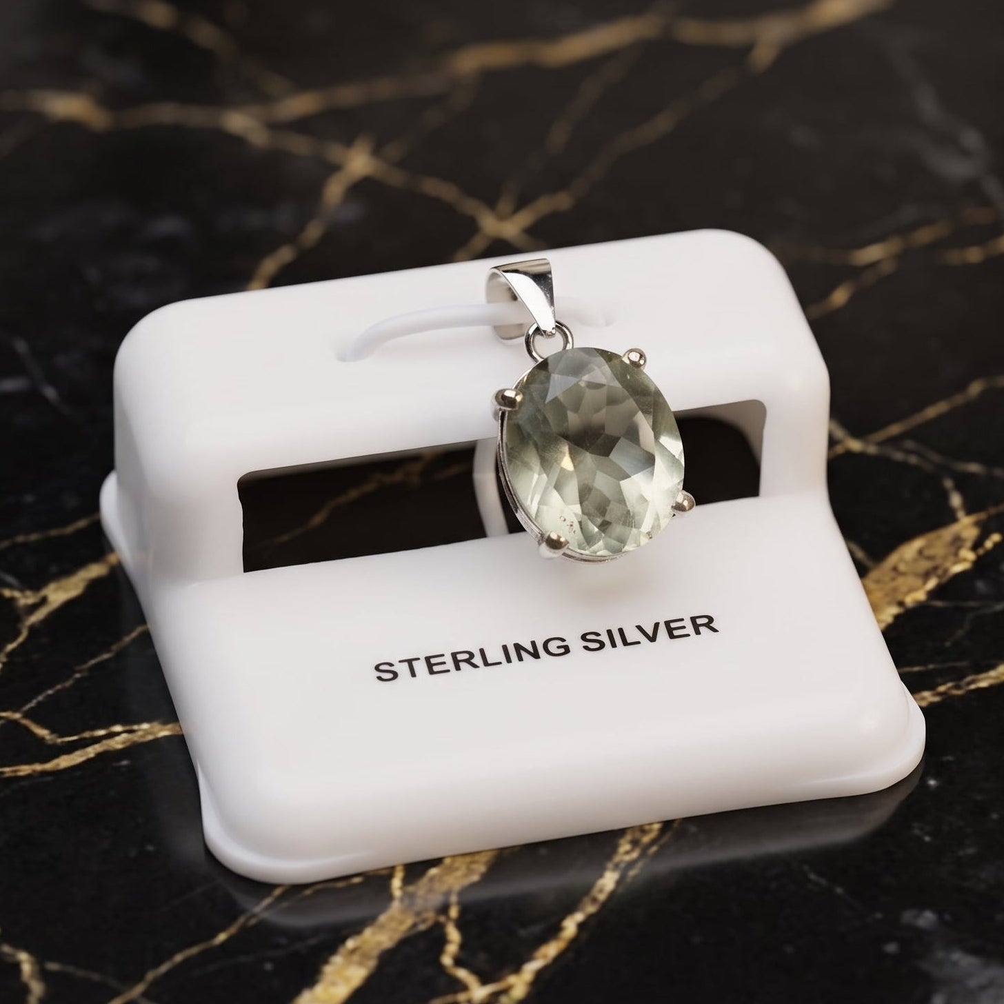 An oval-cut clear gemstone pendant in a sterling silver setting, displayed on a white plastic jewelry stand labeled 'STERLING SILVER' on a black marble surface with gold veining. Green Amethyst