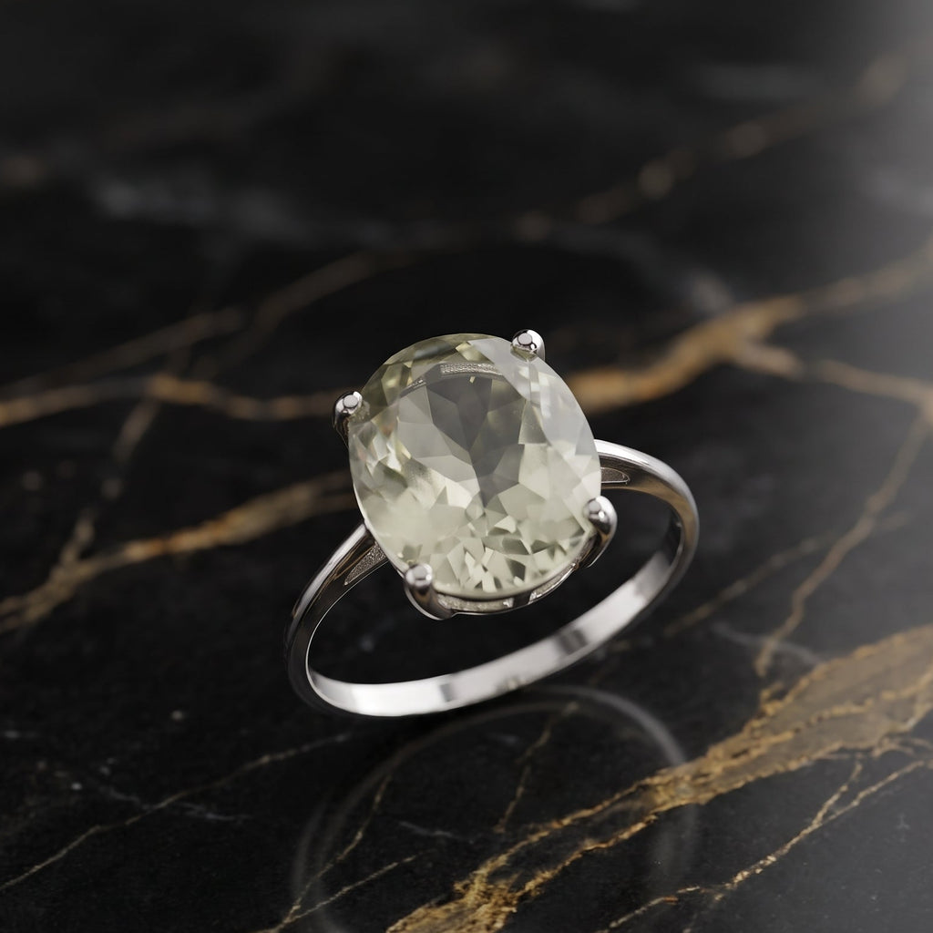 A silver ring with a large oval-cut pale green gemstone, displayed on a black and gold veined marble surface. GREEN AMethyst