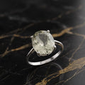 A silver ring with a large oval-cut pale green gemstone, displayed on a black and gold veined marble surface. GREEN AMethyst