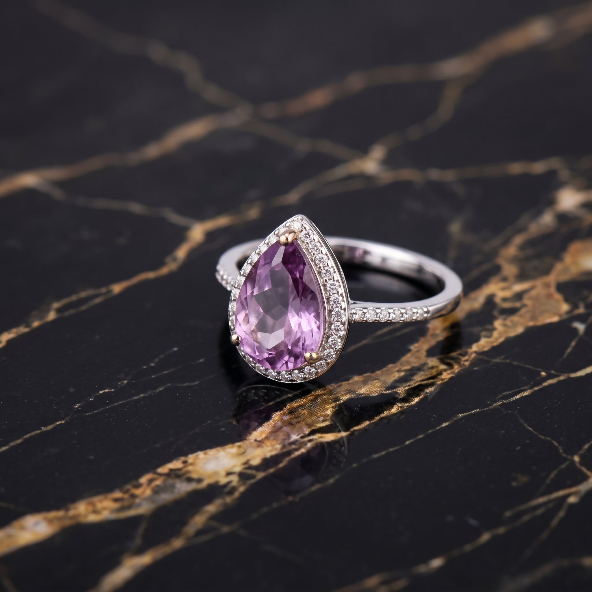 A silver ring featuring a teardrop-shaped pinkish-purple gemstone surrounded by small clear stones, displayed on a black marble surface with gold veining. Amethyst
