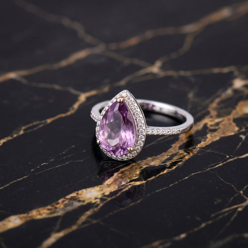 A silver ring featuring a teardrop-shaped pinkish-purple gemstone surrounded by small clear stones, displayed on a black marble surface with gold veining. Amethyst
