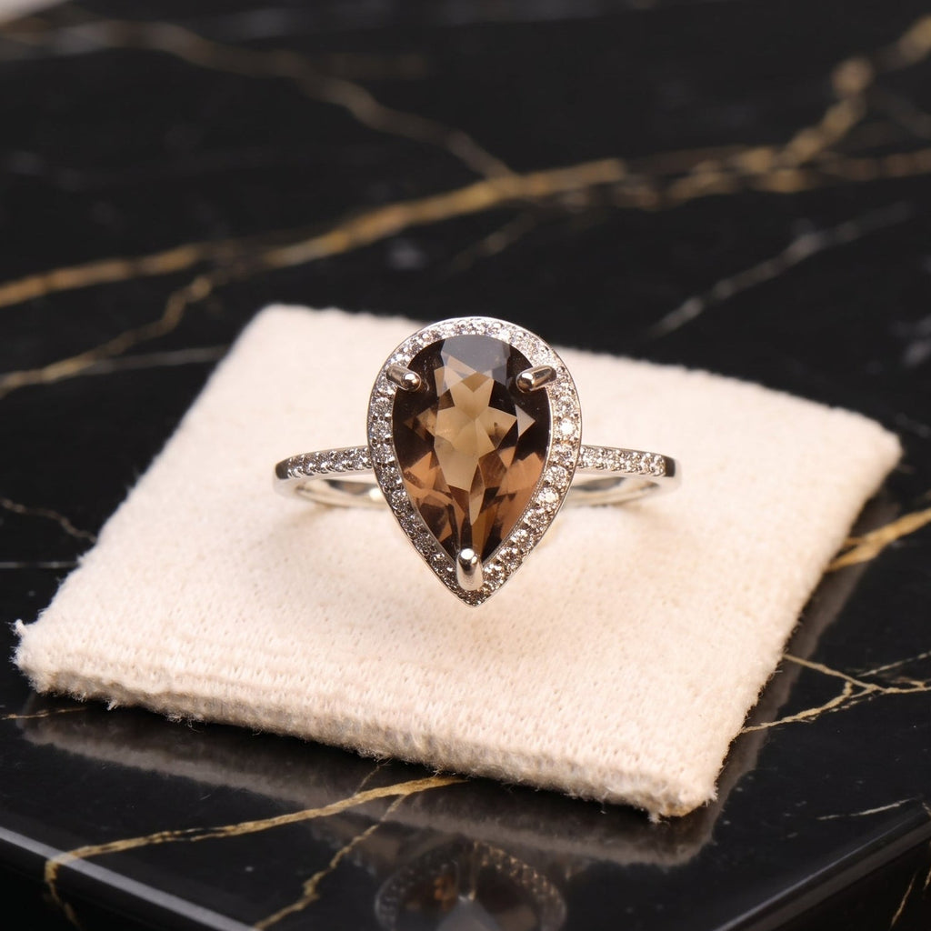 A pear-shaped halo ring with a brown smoky center stone and a silver-toned pavé band, displayed on a cream velvet jewelry cushion atop a black marble surface.