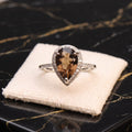A pear-shaped halo ring with a brown smoky center stone and a silver-toned pavé band, displayed on a cream velvet jewelry cushion atop a black marble surface.