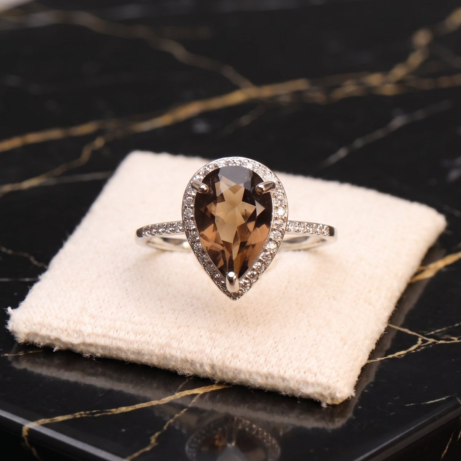 A pear-shaped halo ring with a brown smoky center stone and a silver-toned pavé band, displayed on a cream velvet jewelry cushion atop a black marble surface.