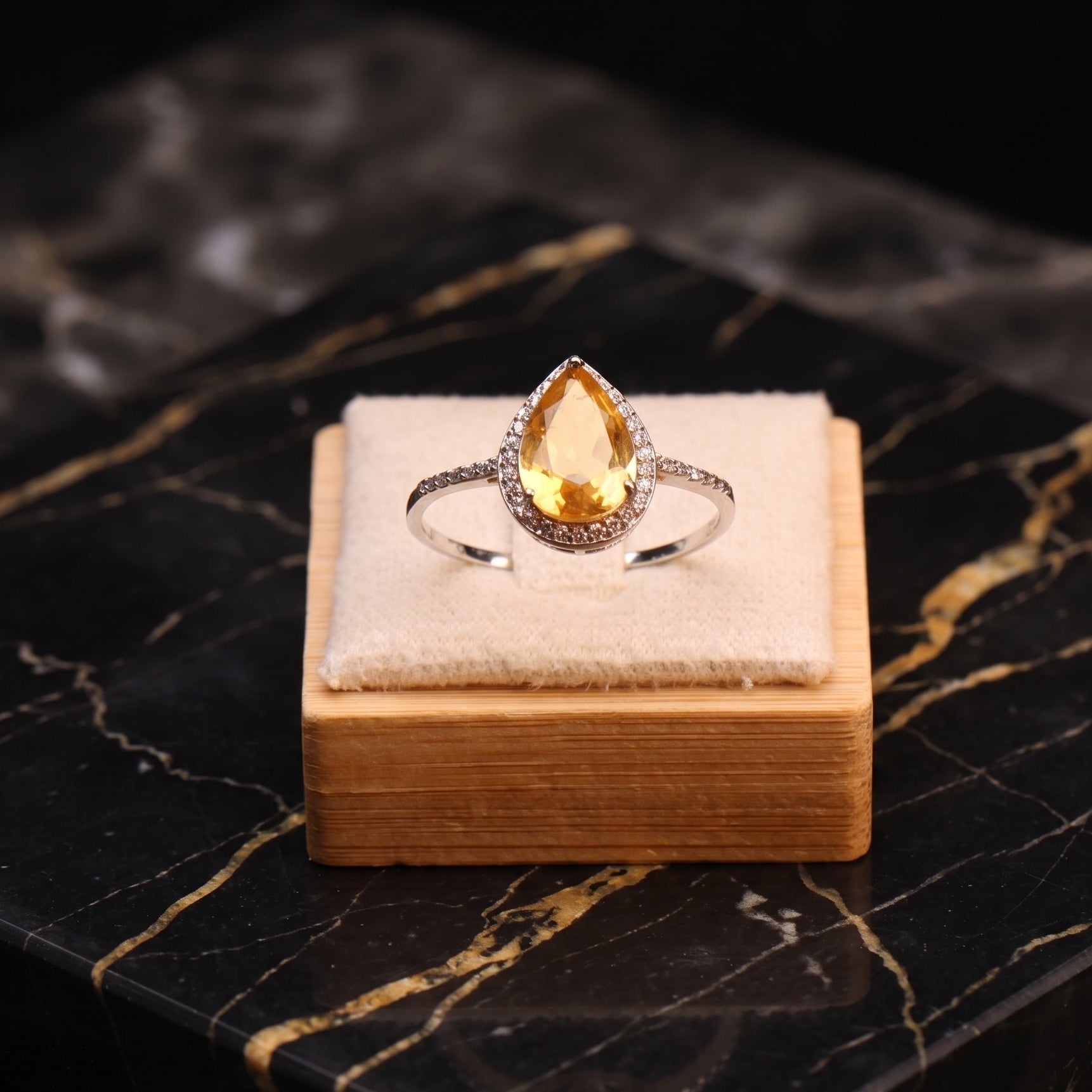 A silver ring with a teardrop-shaped yellow gemstone and small side stones, displayed on a small wooden block with a cream fabric cushion over a black marble surface.