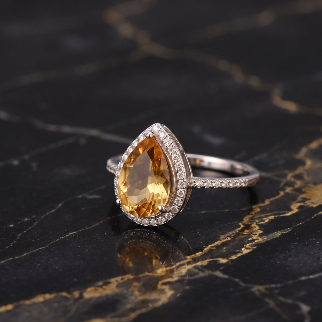 A pear-shaped yellow citrine gemstone ring set in a silver-colored halo with small clear stones on the band, displayed on a black marble surface with gold veining.