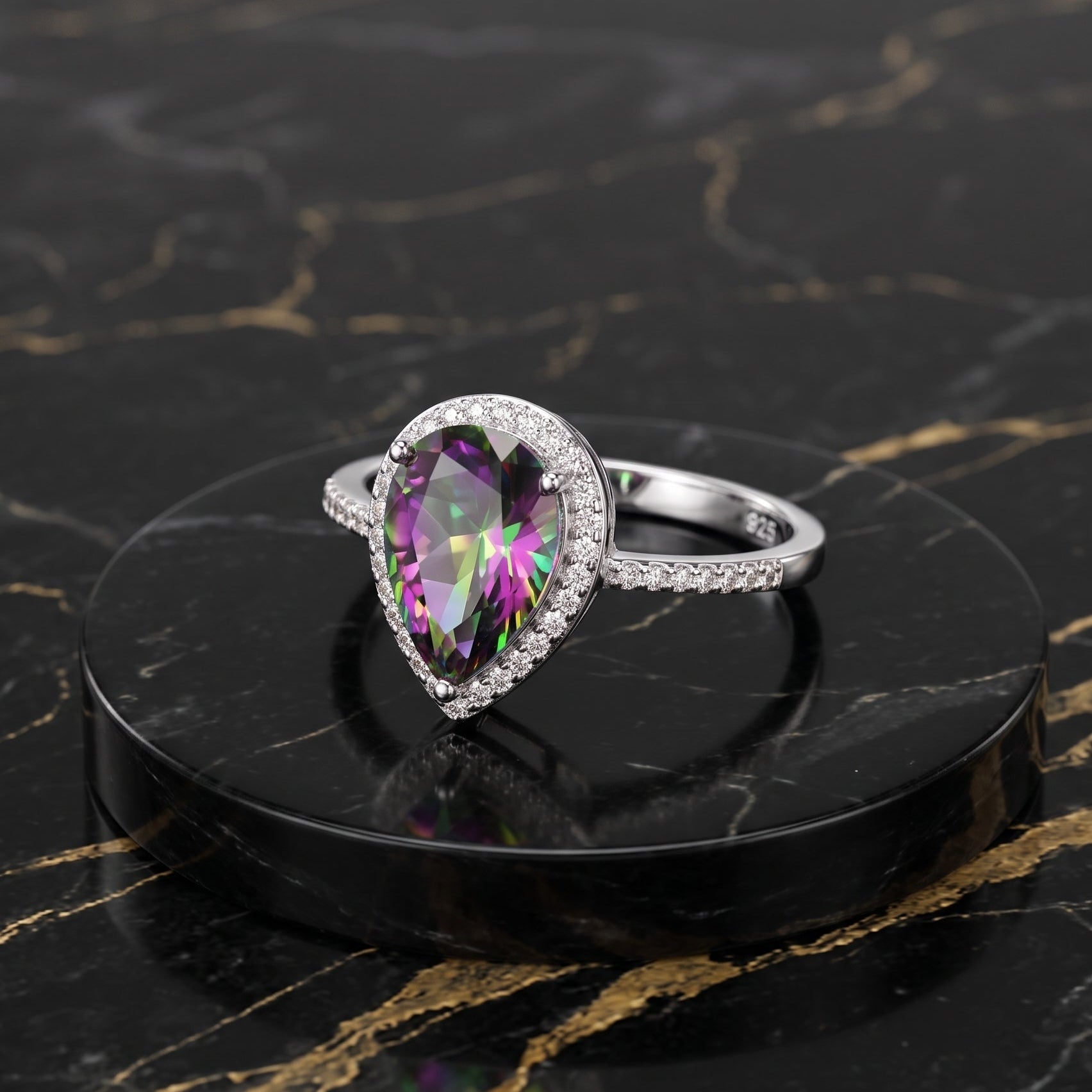 A silver ring with a large teardrop-shaped iridescent purple gemstone and a halo of small clear stones, displayed on a round black marble pedestal over a black marble surface with gold veining. mystic topaz