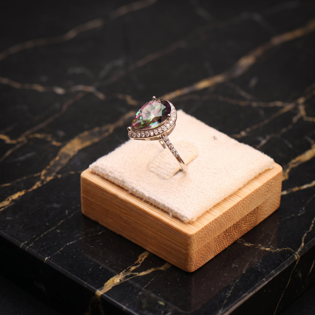 A pear-cut iridescent gemstone ring with a halo of small clear stones on a silver-toned band, displayed on a cream velvet cushion atop a light wood box against a black marble surface.