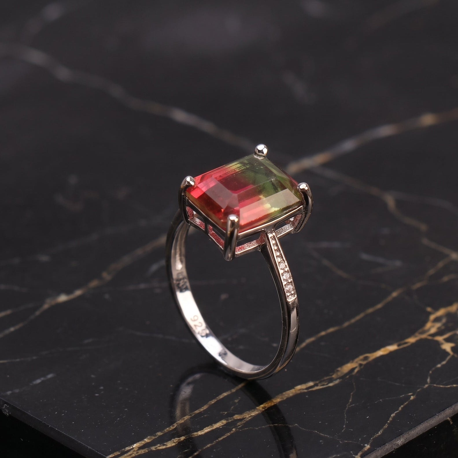 A silver ring featuring a rectangular cushion-cut gemstone with a gradient of red, orange, and green, displayed on a black marble slab with gold veining. watermelon