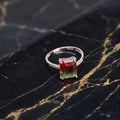 A silver ring featuring a rectangular gradient gemstone that transitions from red at the top to green at the bottom, displayed on a black marble surface with gold veining.