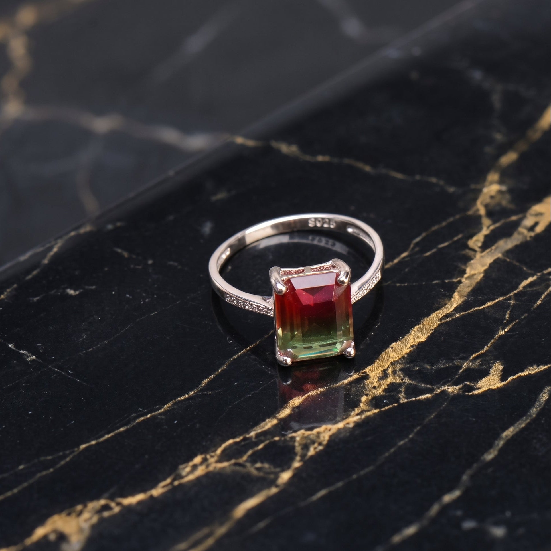 A silver ring featuring a rectangular gradient gemstone that transitions from red at the top to green at the bottom, displayed on a black marble surface with gold veining.