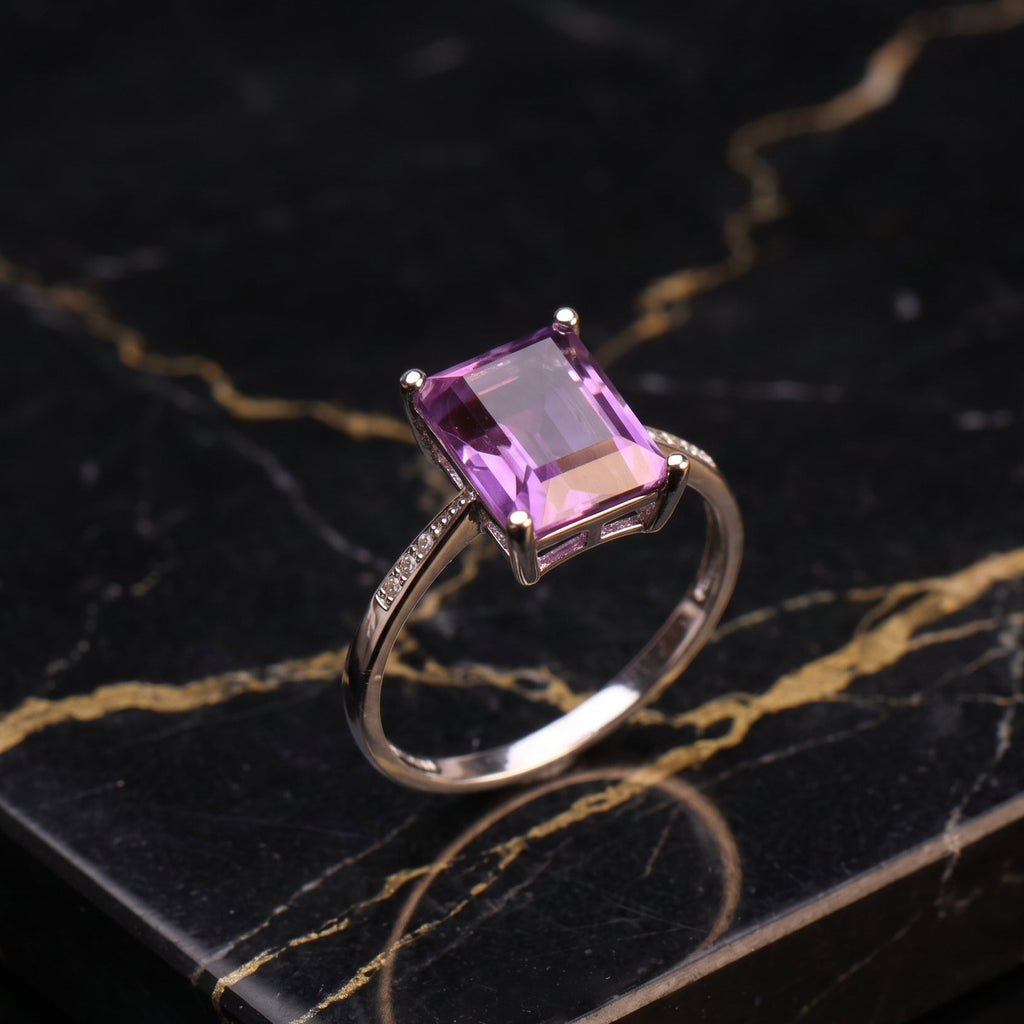 A rectangular purple amethyst gemstone ring set in a silver-colored band, displayed on a black marble slab with gold veining.