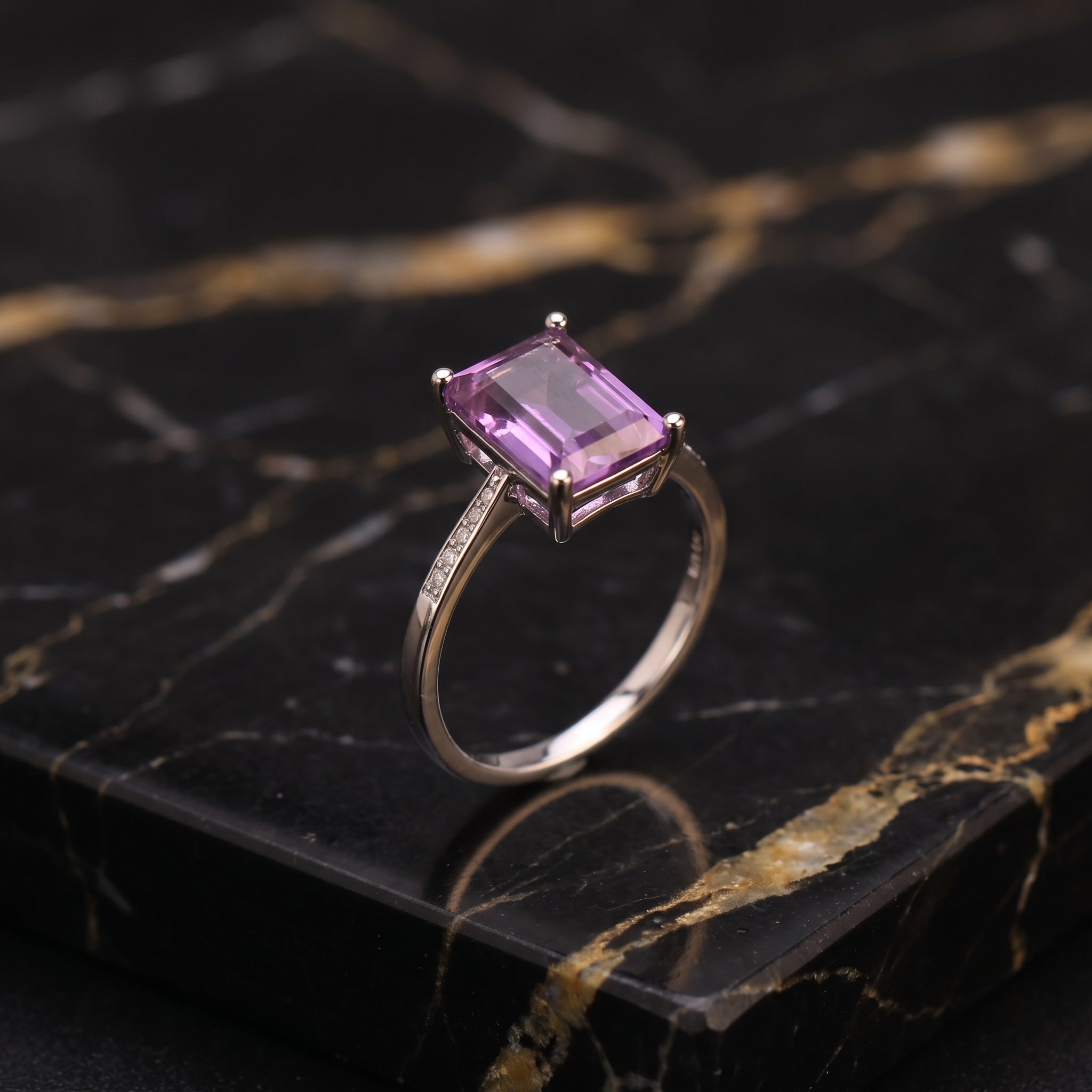 A silver-toned ring with a rectangular purple gemstone, displayed on a black marble slab with gold veining.
