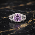 A silver ring featuring a round purple amethyst center stone surrounded by a halo of small clear stones, set on a polished black and gold marble surface.