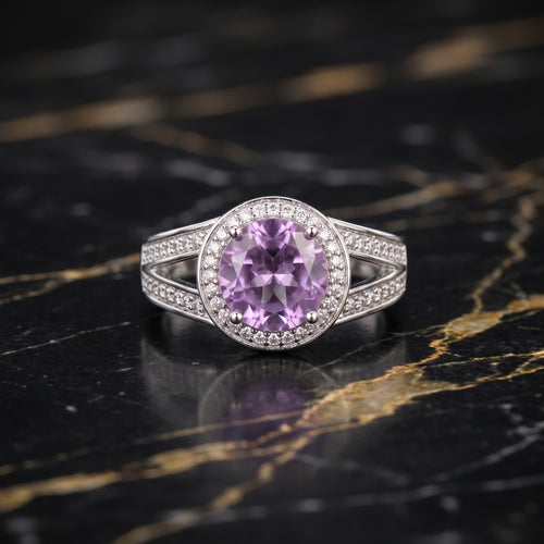 A silver ring featuring a round purple amethyst center stone surrounded by a halo of small clear stones, set on a polished black and gold marble surface.