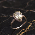 A pear-shaped halo ring featuring a large light green gemstone and small accent stones on a polished silver-toned band, displayed on a black marble surface with gold veining. Green Amethyst