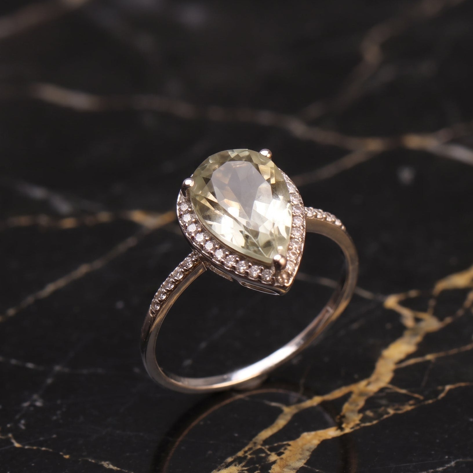A pear-shaped halo ring featuring a large light green gemstone and small accent stones on a polished silver-toned band, displayed on a black marble surface with gold veining. Green Amethyst