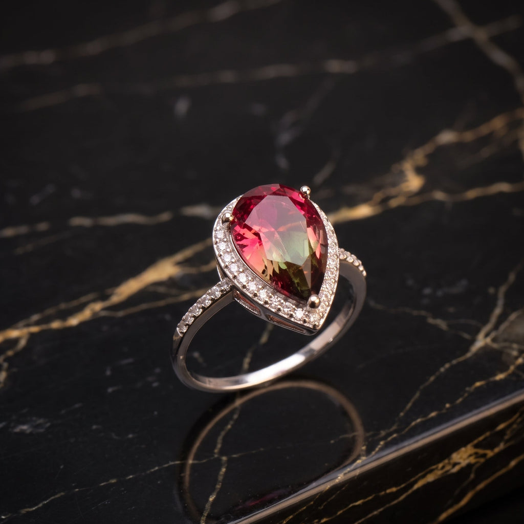 A teardrop-shaped ring featuring a vibrant pinkish-red gemstone in a silver-toned setting with small clear accent stones, displayed on a polished black marble surface with gold veining. watermelon 
