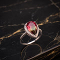 A teardrop-shaped ring featuring a vibrant pinkish-red gemstone in a silver-toned setting with small clear accent stones, displayed on a polished black marble surface with gold veining. watermelon 
