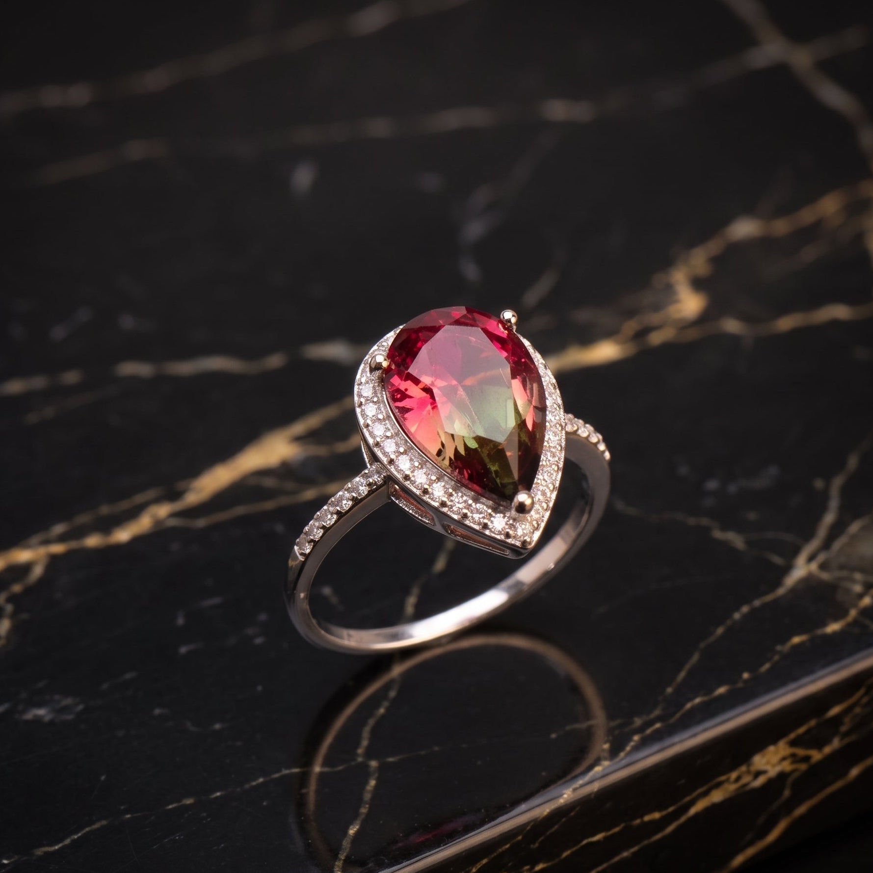 A teardrop-shaped ring featuring a vibrant pinkish-red gemstone in a silver-toned setting with small clear accent stones, displayed on a polished black marble surface with gold veining. watermelon 
