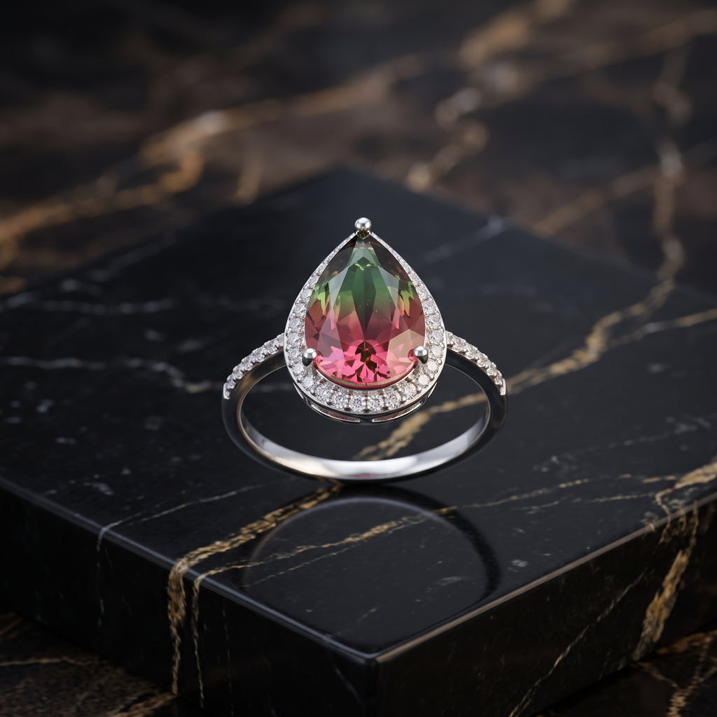 A teardrop-shaped ring with a faceted pink and green gemstone, surrounded by a halo of small clear stones and a polished silver band, displayed on a black marble block. Watermelon