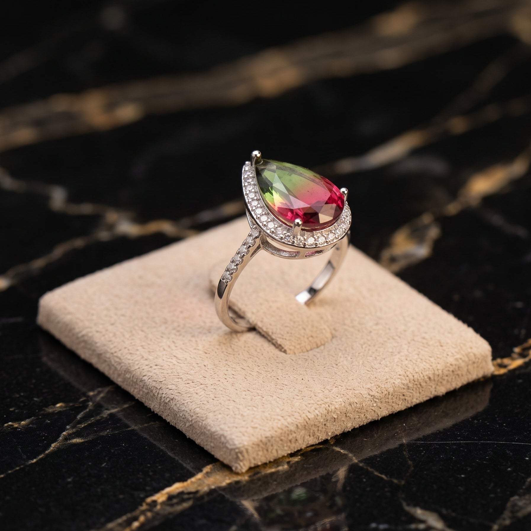 A silver ring with a large teardrop-shaped multicolor gemstone featuring green, pink, and yellow hues, displayed on a beige velvet ring stand over a black marble surface.