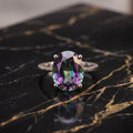 An oval-cut iridescent gemstone ring with a silver-toned band, resting on a polished black marble surface with gold veining. mystic topaz