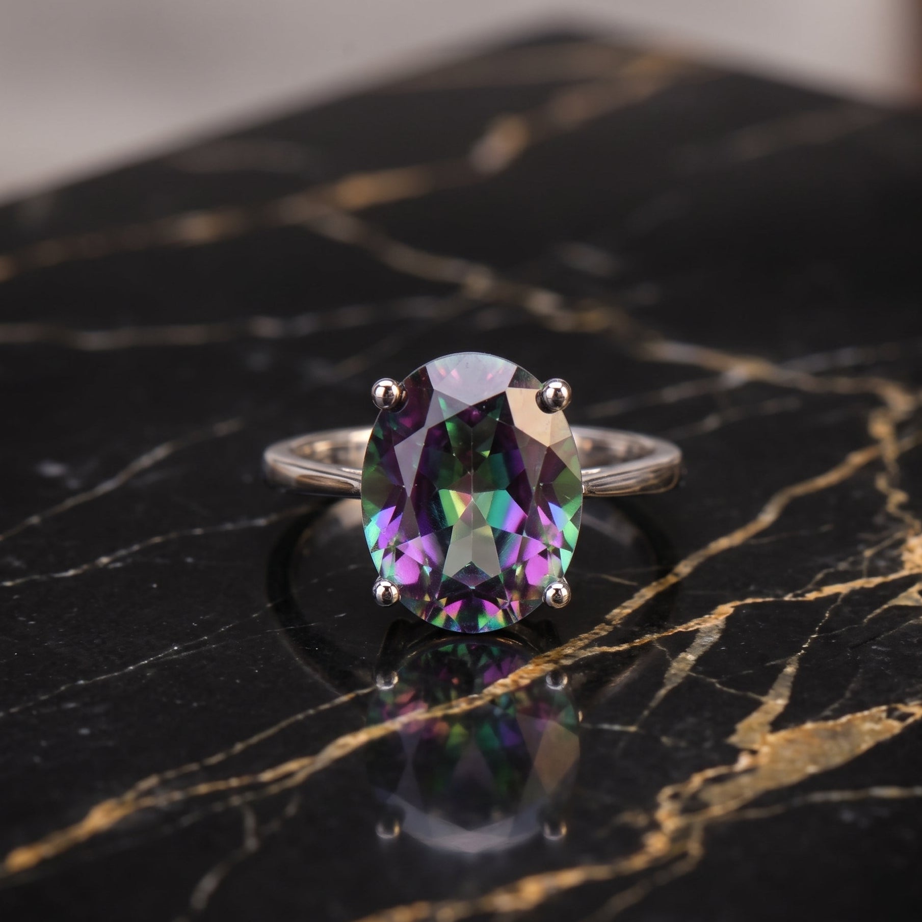 An oval-cut iridescent gemstone ring with a silver-toned band, resting on a polished black marble surface with gold veining. mystic topaz