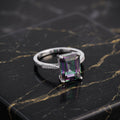 A silver ring featuring a rectangular emerald-cut iridescent gemstone with purple, green, and blue color shifts, displayed on a black marble slab with gold veining. mystic topaz
