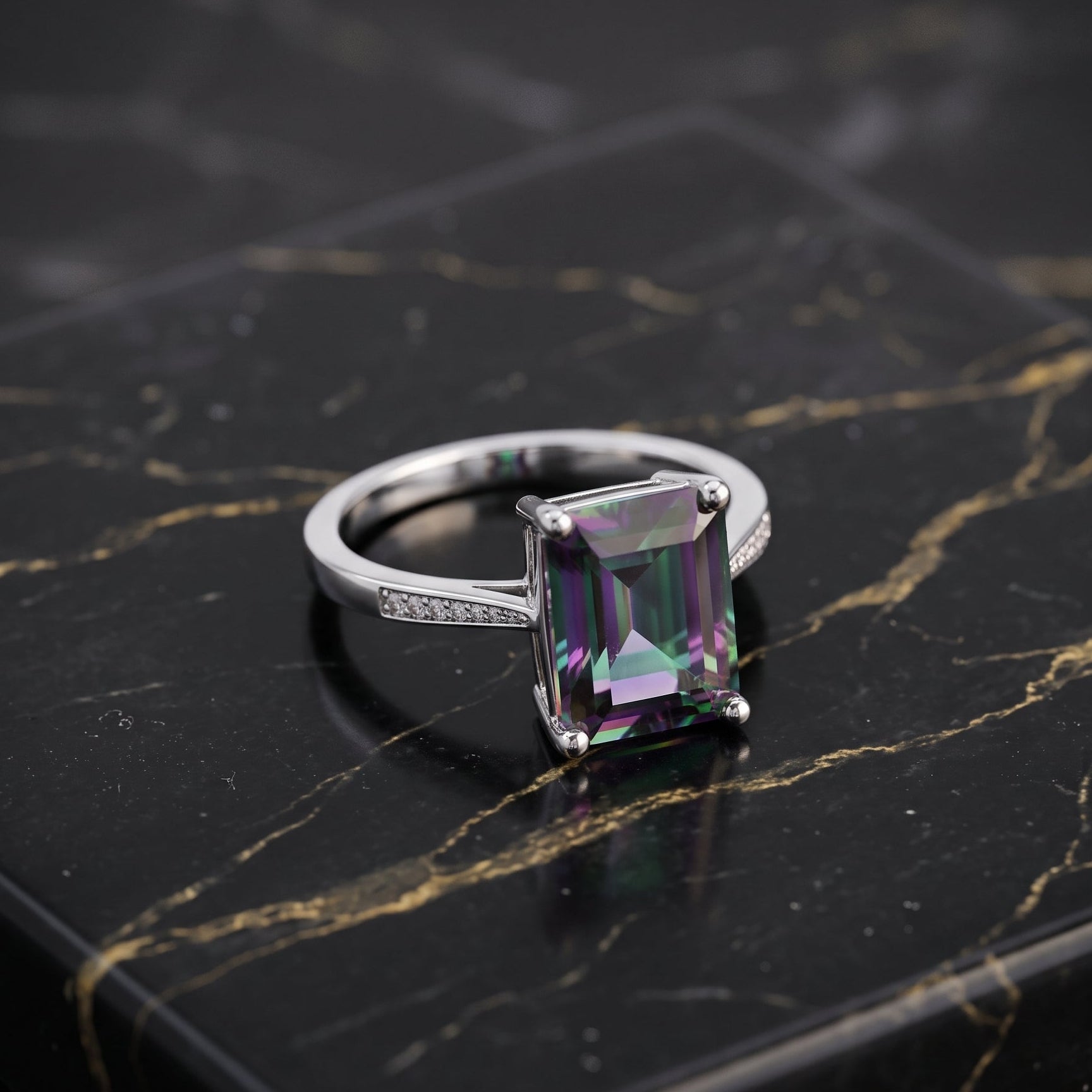 A silver ring featuring a rectangular emerald-cut iridescent gemstone with purple, green, and blue color shifts, displayed on a black marble slab with gold veining. mystic topaz
