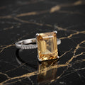 A silver ring featuring a large rectangular emerald-cut yellow gemstone, displayed on a black marble surface with gold veining. citrine