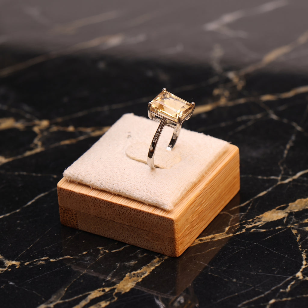 A yellow emerald-cut gemstone ring on a silver band, displayed on a small wooden stand with a white velvet cushion, set against a black marble background with gold veining.