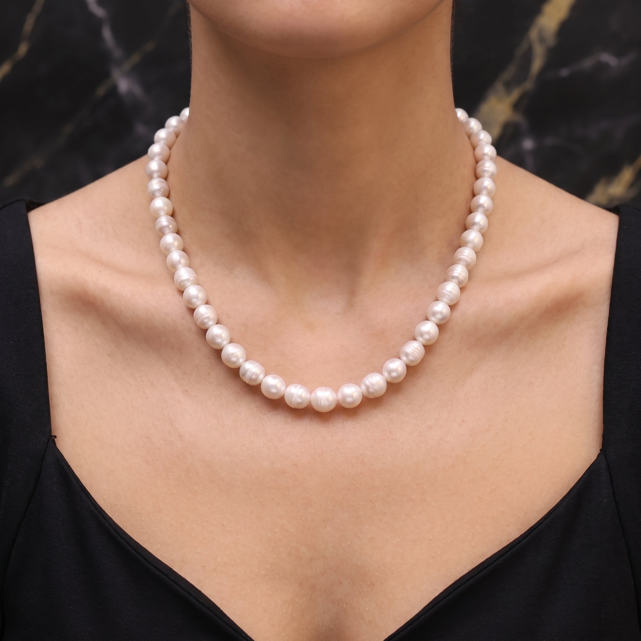 A close-up of a model wearing a classic single-strand pearl necklace with white, lustrous pearls, paired with a black top against a dark marble background.