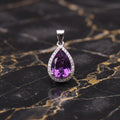 A teardrop-shaped purple amethyst pendant set in a silver-colored halo with small clear stones, displayed on a black and gold marbled surface.