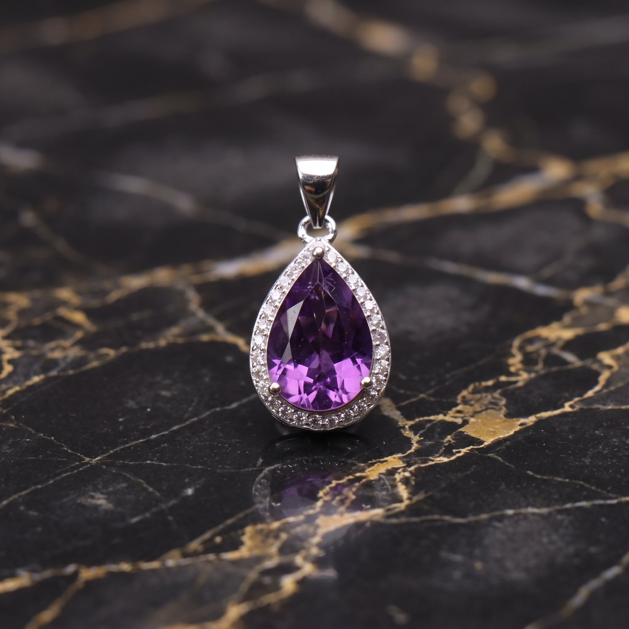 A teardrop-shaped purple amethyst pendant set in a silver-colored halo with small clear stones, displayed on a black and gold marbled surface.